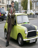 A man dressed as Mr. Bean leans against a lime green classic Mini car on a city street.