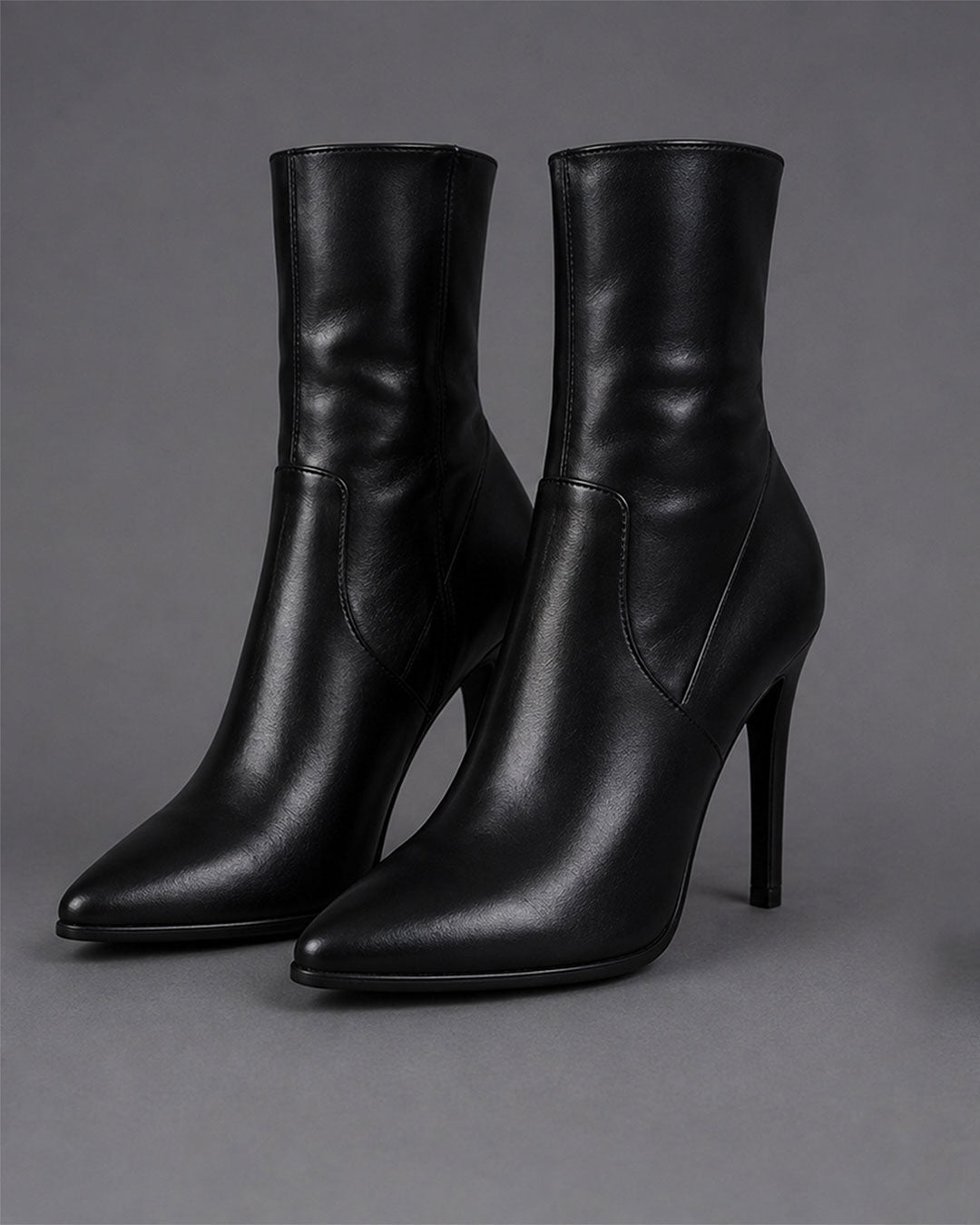 A pair of black leather ankle boots with pointed toes and slim high stiletto heels, photographed on a neutral gray studio background.