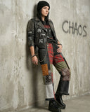 Person wearing a punk-style outfit with studded jacket and patchwork pants against a concrete wall with 'CHAOS' graffiti.