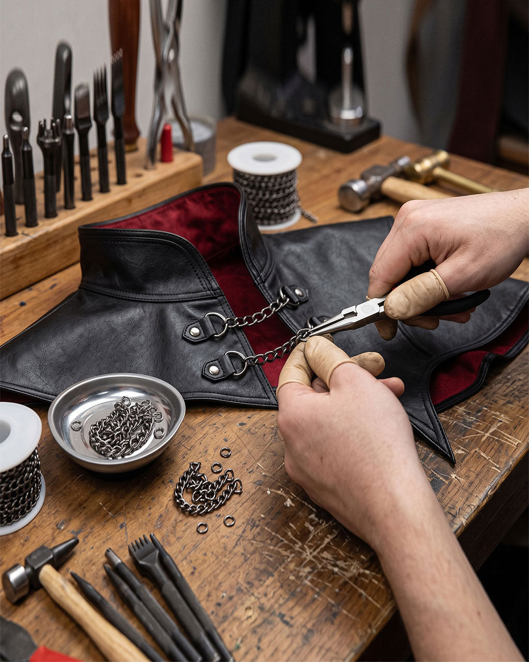 A close-up of a black leather jacket or vest with a red lining, being assembled on a wooden workbench. Hands are shown attaching a metal chain with pliers, surrounded by leatherworking tools and chains.